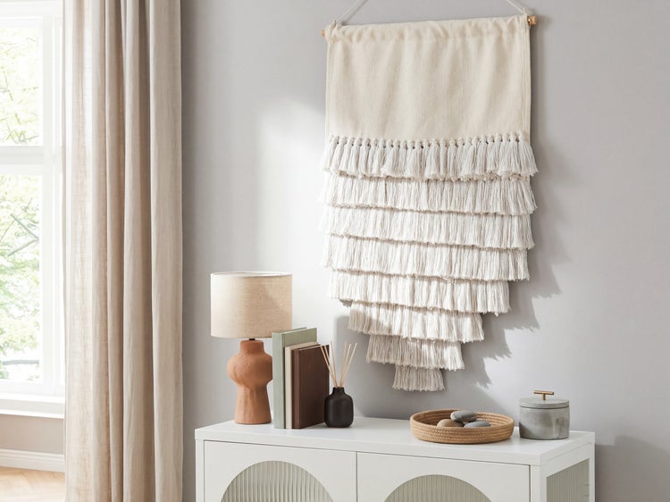 Textiler Wandbehang mit mehreren Reihen Fransen über einem weißen Sideboard mit Lampe und Dekoration in einem hellen Raum.
