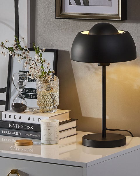 Moderne schwarze Tischleuchte mit halbkugelförmigem Schirm auf einer Kommode neben Büchern, einer Vase mit Blumen und einer Kerze in einem hellen Raum.