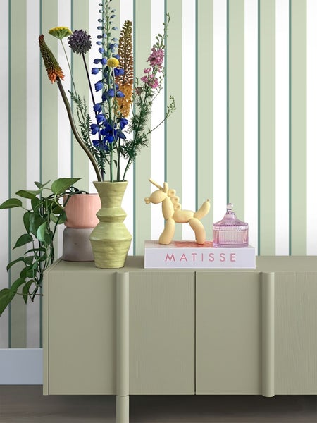 Dekoratives Arrangement mit Vase, Blumen und Zierobjekten auf einem Schrank vor einer gestreiften Tapete.