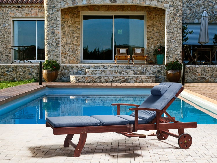 Holz-Sonnenliege mit blauer Auflage und Rollen am Pool vor einem Natursteinhaus auf einer hellen Terrasse.