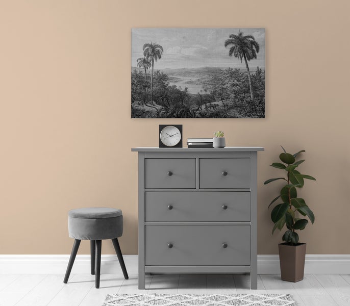 Wohnraum mit grauer Kommode, gepolstertem Hocker, Wandbild mit Palmenmotiv, Tischuhr, Büchern und einer Zimmerpflanze in einem Topf.
