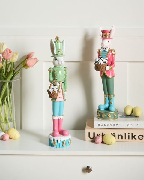 Osterdekoration mit zwei Hasenfiguren im Nussknacker-Stil, Tulpen in einer Vase und Deko-Eiern auf einer weißen Ablage.