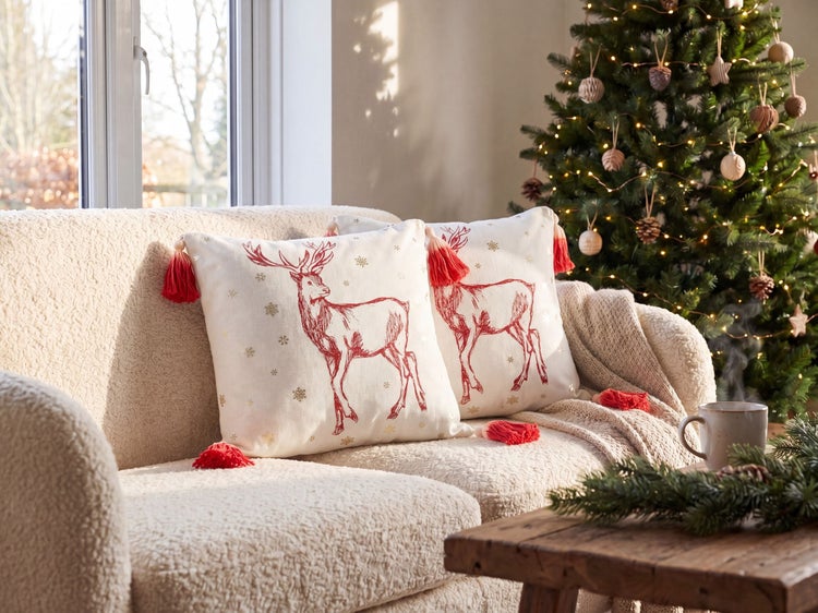 Dekokissen mit Hirsch-Stickerei und Quasten auf einem Sofa in einem hellen Wohnzimmer mit festlich geschmücktem Weihnachtsbaum.