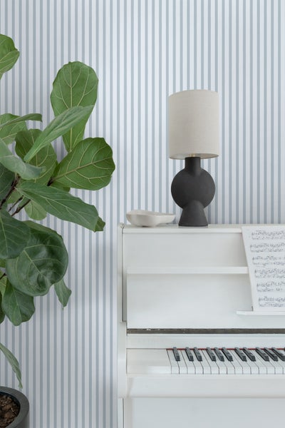 Dekoratives Interieur mit Zimmerpflanze, Lampe und weißem Klavier vor gestreifter Tapete