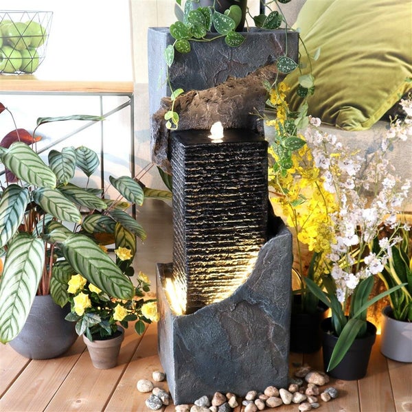 Dekorativer Zimmerbrunnen mit Beleuchtung und Steinoptik, umgeben von Zimmerpflanzen auf einem Holzboden.