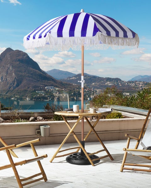 Blau-weiß gestreifter Sonnenschirm mit Fransen auf einer Terrasse mit Bambusmöbeln und Bergblick.