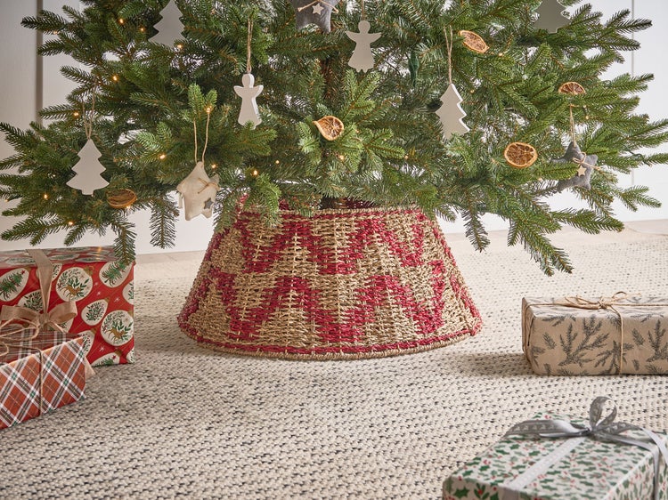 Weihnachtsbaum mit Baumschmuck und geflochtenem Baumständer sowie Geschenken auf einem Teppich