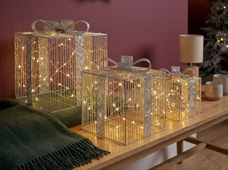 Dreier-Set dekorative LED-Geschenkboxen in verschiedenen Größen aus Metalldraht mit warmweißer Beleuchtung auf einer Holzbank.