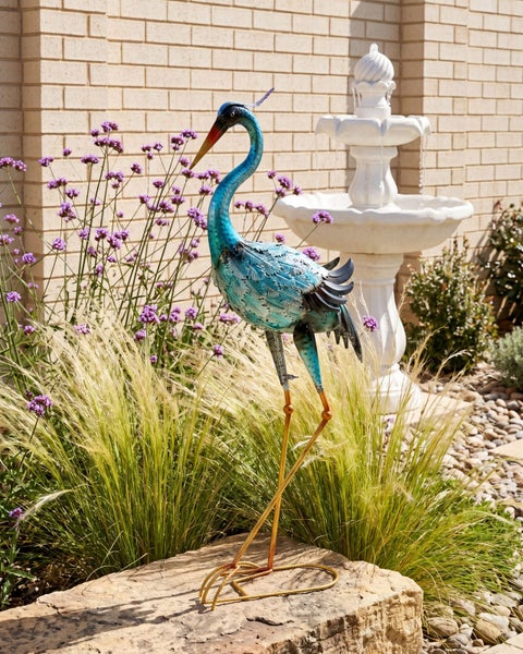 Gartenfigur Kranich aus Metall in einem Garten mit Gräsern, Blumen und einem Springbrunnen.