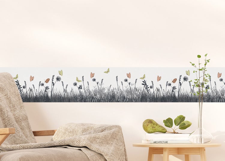 Tapetenbordüre mit Wiesenmotiv und Schmetterlingen an einer Wand über einem Sessel und Beistelltisch in einem hellen Wohnzimmer.