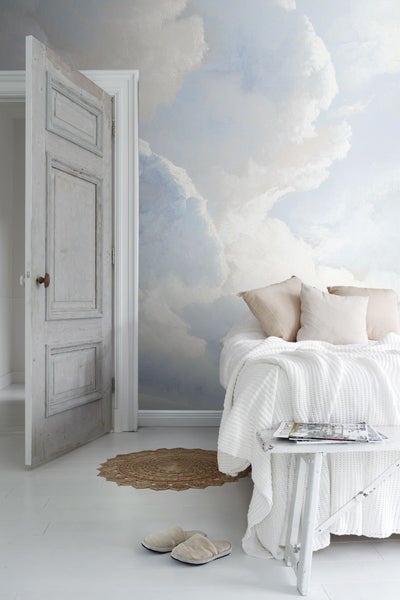 Helles Zimmer mit Wolken-Tapete, Bett, Beistelltisch und geöffneter Holztür