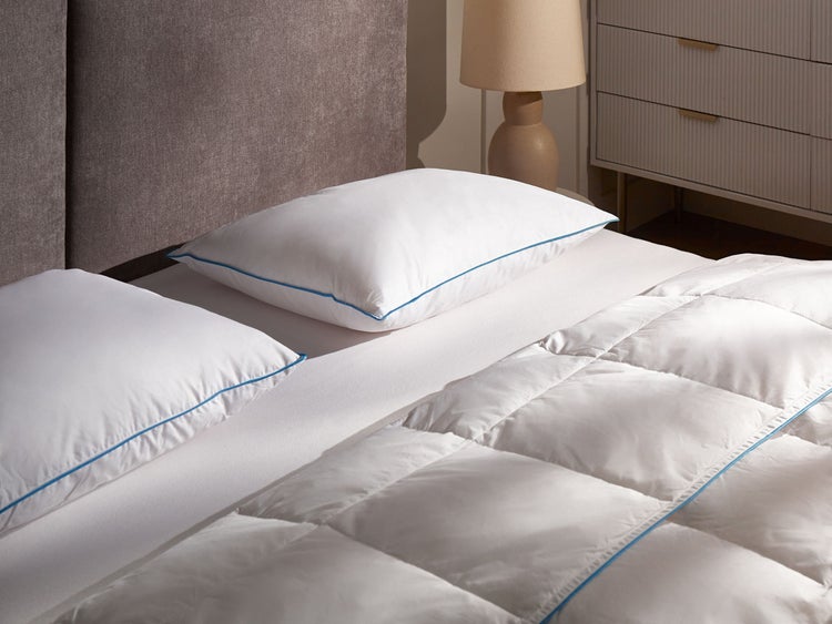 Weißes Bettwäsche-Set mit zwei Kopfkissen und Steppdecke mit blauem Rand auf einem Bett mit grauem Kopfteil.
