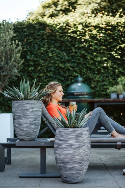 Zwei strukturierte hohe Pflanzgefäße mit Agaven auf einer Terrasse neben einer Gartenliege und einem Keramikgrill vor einer Hecke.