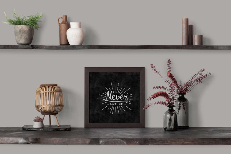 Quadratisches Wandbild mit dem Schriftzug Never Give Up in dunklem Rahmen auf einem Holzregal neben Windlicht und Vasen mit Trockenblumen.