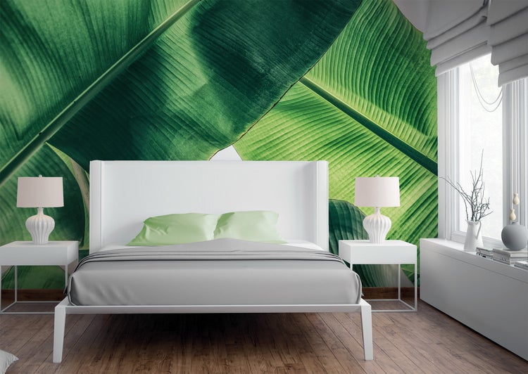 Helles, modernes Schlafzimmer mit großflächiger Bananenblatt-Fototapete, weißem Bett mit hellgrünen Kissen, weißen Nachttischen und Holzboden.