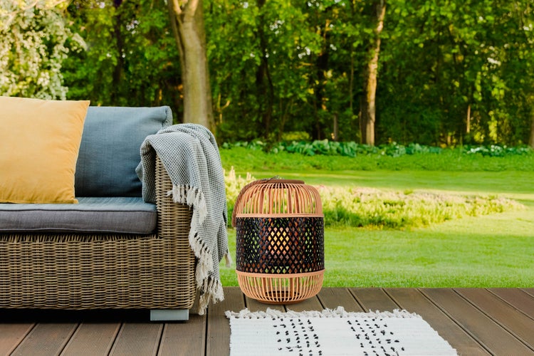 Szene mit Rattansofa, Kissen, Decke und Bambuslaterne auf einer Holzterrasse im Garten.