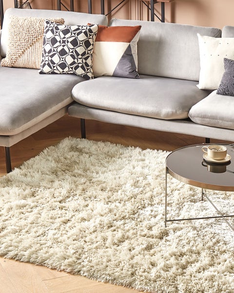 Wohnzimmer mit grauem Ecksofa, dekorativen Kissen, flauschigem Hochflorteppich und rundem Beistelltisch auf einem Holzboden.