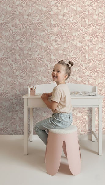 Helles Kinderzimmer mit Zebramuster-Vliestapete an der Wand, weißem Schreibtisch und einem Kind auf einem Hocker.