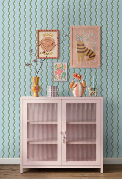 Hellrosa Vitrine mit geriffelten Glastüren vor einer Wand mit grünem Wellenmuster und verschiedenen Dekorationsgegenständen.