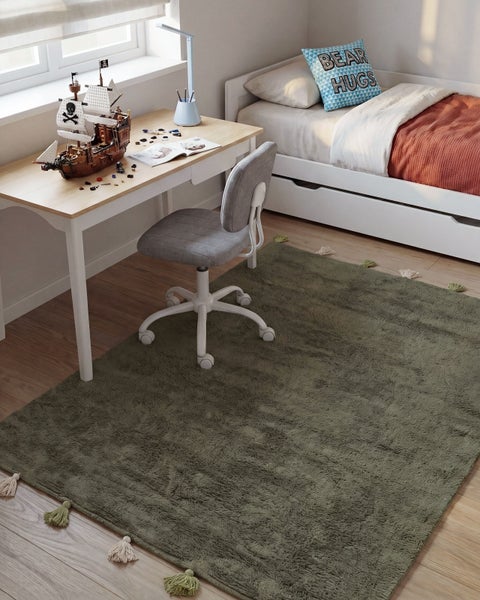 Helles Kinderzimmer mit Holztisch, Schreibtischstuhl, Einzelbett und einem flauschigen Teppich mit Quasten auf dem Parkettboden.