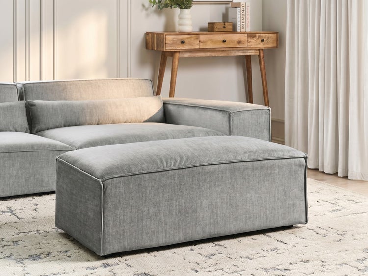 Modernes Wohnzimmer mit einem großen rechteckigen Polsterhocker und passendem Ecksofa auf einem gemusterten Teppich vor einer Konsole aus Holz.