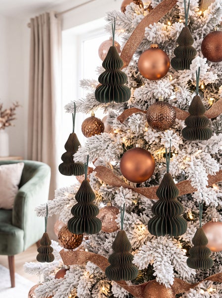 Weihnachtsbaum mit Kunstschnee, geschmückt mit Kugeln und tannenförmigen Papieranhängern in einem hellen Wohnzimmer.