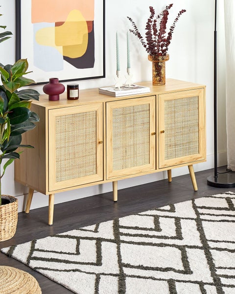 Sideboard aus hellem Holz mit drei Türen und Rattangeflecht, auf konischen Beinen in einem hellen Wohnzimmer mit geometrischem Teppich.