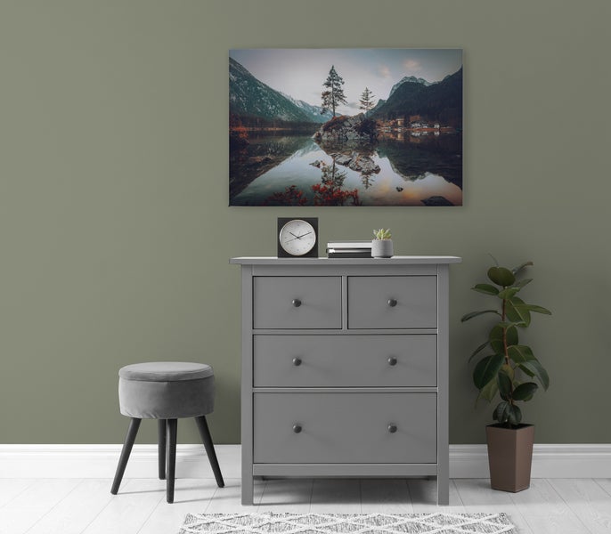 Wohnraum mit einer grauen Kommode, einem Hocker, einer Zimmerpflanze und einem Wandbild mit Bergsee am Hintergrund.