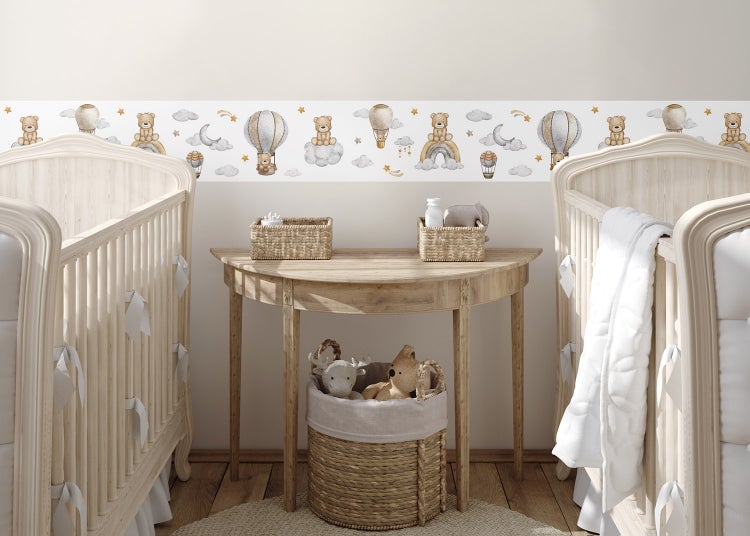 Wandbordüre mit Motiven von Teddybären und Heißluftballons in einem hellen Kinderzimmer mit Holztisch und Gitterbetten.