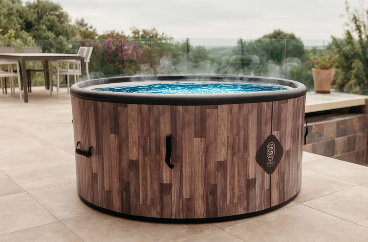 Runder aufblasbarer Whirlpool in Holzoptik mit COCO SPA Logo und dampfendem Wasser auf einer gefliesten Terrasse im Garten.