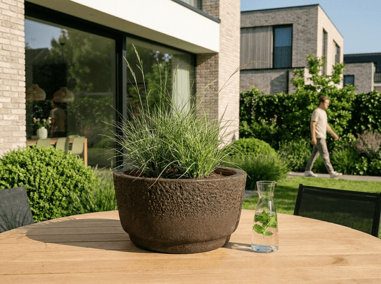 Runder Pflanztopf mit strukturierter Oberfläche und dekorativem Gras auf einem Holztisch im Garten vor einem modernen Wohnhaus.