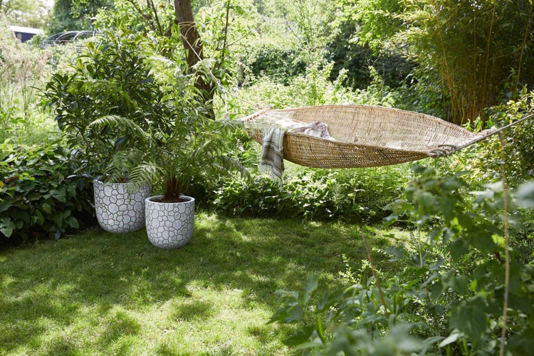 Ein sonniger Garten mit schiffchenförmiger Korb-Hängematte zwischen Bäumen und zwei großen gemusterten Pflanztöpfen auf einer Rasenfläche.