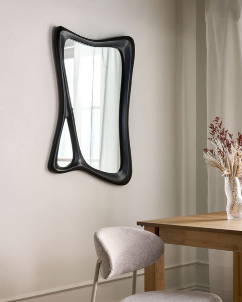 Unregelmäßiger Spiegel mit schwarzem Rahmen über einem Holztisch und einem Stuhl