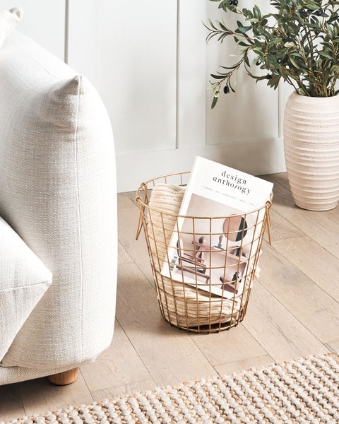 Eine helle und gemütliche Wohnzene mit einem Sofa, einem Drahtkorb mit Decke und Zeitschrift sowie einem Dekozweig in einer Vase.