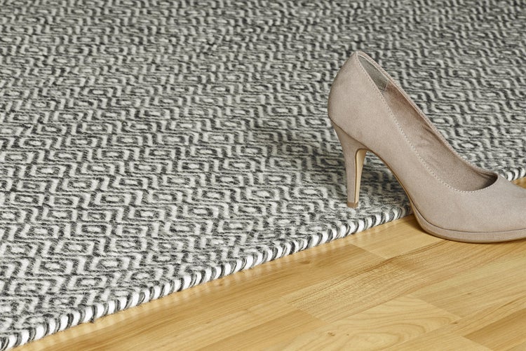 Flachgewebter Teppich mit grauweißem geometrischem Muster auf Holzfußboden mit einem beigen Damenschuh.