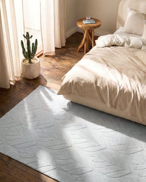 Hellblauer Teppich mit Blattmuster in einem Schlafzimmer auf dunklem Holzboden, neben einem Bett und einer Kaktee.