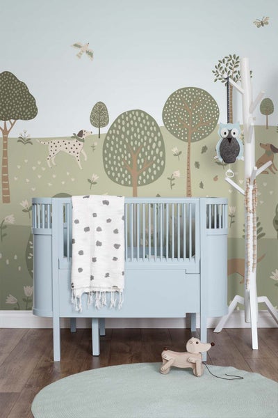 Kinderzimmer mit hellblauem Babybett und Vliestapete mit Waldmotiven wie Bäumen, Hunden und Vögeln im skandinavischen Design.