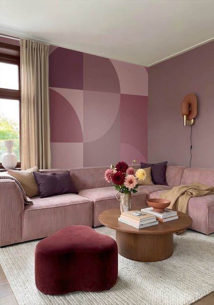 Modernes Wohnzimmer mit Ecksofa aus Cord, geometrischer Wandgestaltung, rundem Couchtisch aus Holz und einem Samt-Hocker auf hellem Teppich.
