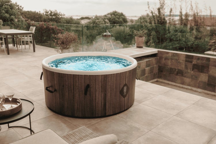 Runder Außenwhirlpool in Holzoptik auf einer gefliesten Terrasse mit Glasgeländer und Essbereich im Hintergrund.