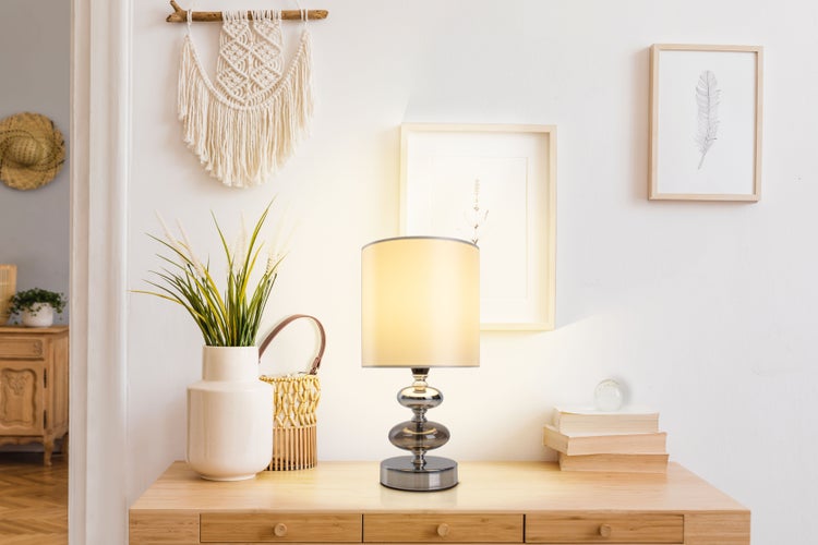 Dekoratives Interieur mit Tischlampe, Vase mit Grünpflanzen, Wandbehang und Bilderrahmen auf einem Holztisch