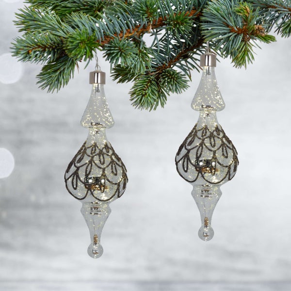 Zwei gläserne Christbaumkugeln mit Ornamenten hängen an einem Tannenzweig.