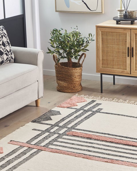 Hellgrundiger Flachgewebe-Teppich mit geometrischem Muster in Schwarz und Rosa sowie Fransen, kombiniert mit modernen Holzmöbeln und einer Pflanze.