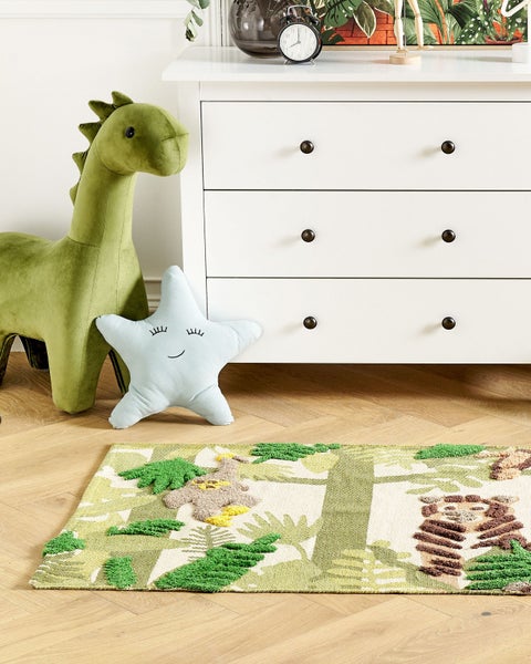 Helles Kinderzimmer mit gemustertem Kinderteppich, weißer Kommode, grünem Plüsch-Dinosaurier und hellblauem Sternenkissen auf Holzboden.