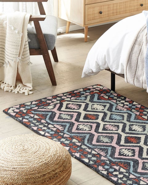 Rechteckiger Teppich mit buntem geometrischem Muster auf hellem Holzboden in einem hellen Schlafzimmer neben Bett, Pouf und Sessel.