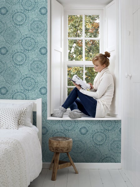 Helles Zimmer mit floral gemusterter Tapete und einer Frau, die in einem Fenster sitzt und liest