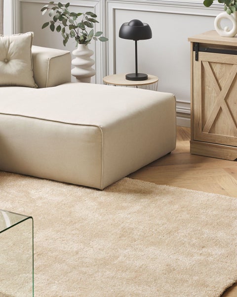 Ein helles Wohnzimmer mit Sofa, Beistelltisch, Stehlampe und Teppich vor einem Holzschrank im Landhausstil auf einem Parkettboden.
