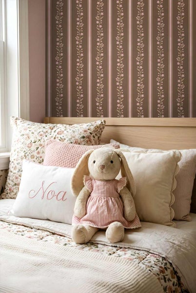 Kinderbett mit hölzernem Kopfteil, verschiedenen Kissen mit floralen Mustern und Bestickungen sowie einem Stoffhasen vor einer gestreiften Tapete mit Rankenmuster.