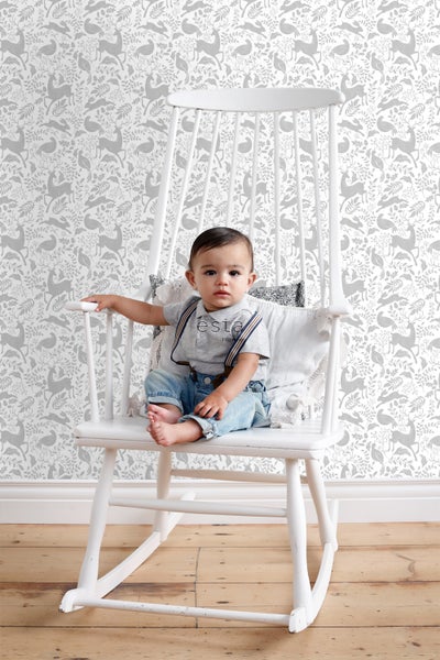 Ein Baby sitzt in einem weißen Schaukelstuhl vor einer Tapete mit Tiermotiven.