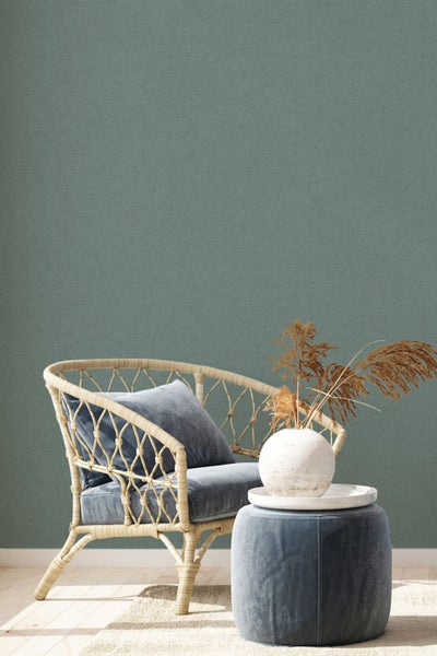 Wohnszene mit Rattansessel, Kissen, Beistelltisch und Vase mit Trockenblumen vor einer einfarbigen Wand