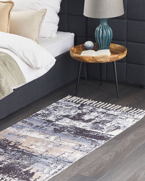 Rechteckiger Teppich mit abstraktem Vintage-Muster in Blau und Grau, weiße Fransen. Im Schlafzimmer auf dunklem Holzboden neben einem Bett.